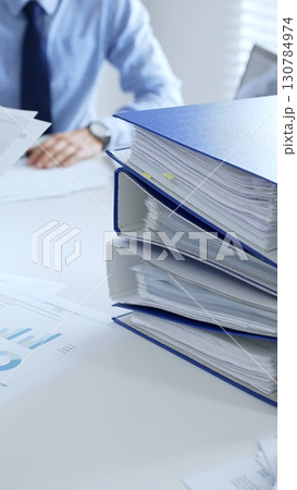 Corporate professionals reviewing financial documents, working collaboratively at white desk with blue folder stack, highlighting teamwork and business productivity. Audit and taxes in business 130784974