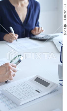 Two accountants analyzing financial reports with magnifying glass and documents, performing accounting and auditing tasks in the office, close up of hands. Audit and taxes in business Two accountants analyzing financial reports with magnifying glass and documents, performing accounting and auditing tasks in the office, close up of hands. Audit and taxes in business 130785094