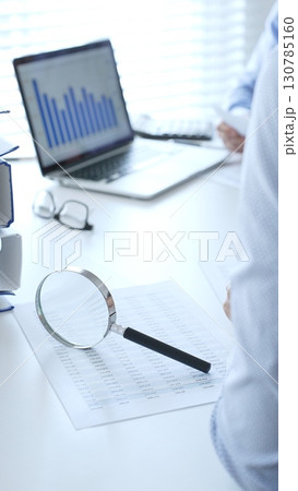 Magnifying glass over financial documents near blue folders full of papers, accountants are analyzing reports and documents at the background. Audit and taxes in business 130785160