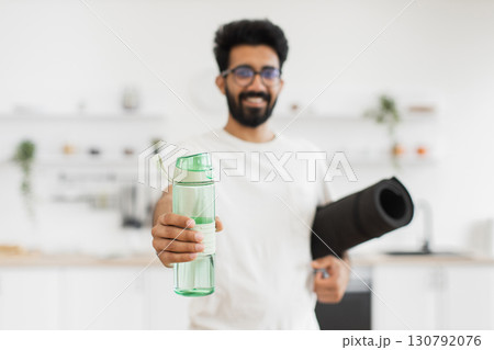 Young man wearing glasses standing in cozy home kitchen holding water bottle and yoga mat. Promotes hydration, comfort, healthy lifestyle, and happiness. 130792076