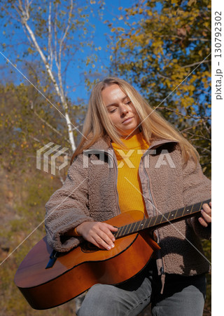 Portrait of smiling caucasian artist plays wooden acoustic guitar in autumnal park. Young songwriter plays string musical instrument hobby outside in nature fall time. Audio music healing Portrait of smiling caucasian artist plays wooden acoustic guitar in autumnal park. Young songwriter plays string musical instrument hobby outside in nature fall time. Audio music healing 130792302