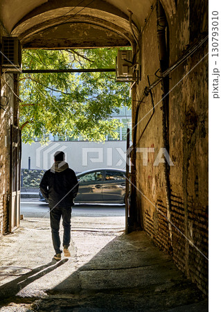 A man walks through a rustic archway, illuminated by a ray of sunlight. The dramatic light and shadow create a contrast, emphasizing the feeling of urban journey. A man walks through a rustic archway, illuminated by a ray of sunlight. The dramatic light and shadow create a contrast, emphasizing the feeling of urban journey. 130793010