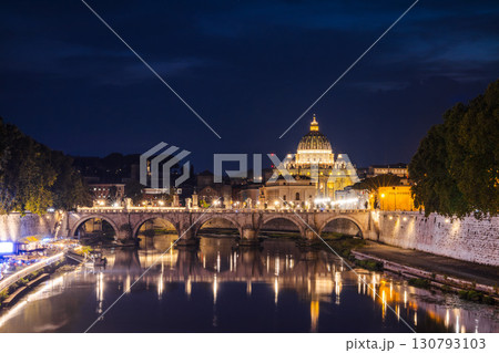 Illuminated St. Peter's Basilica in Vatican City with the glowing Ponte Sant'Angelo bridge reflecting in the Tiber River at night, symbolizing faith, history, and timeless Roman beauty 130793103