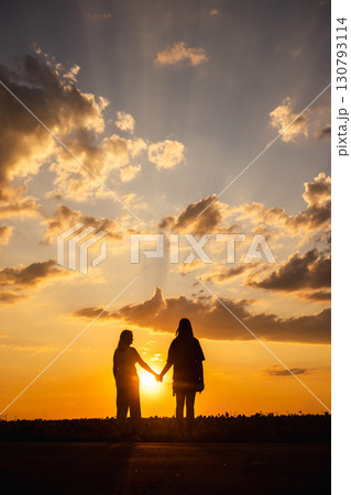 Two women holding hands in silhouette at sunset with dramatic clouds and rays of light in the sky 130793114