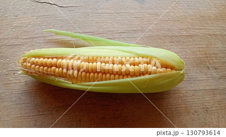 A single ear of ripe corn with its green husk partially peeled back, showcasing vibrant yellow kernels on a rustic wooden background. Captured in a top down view 130793614
