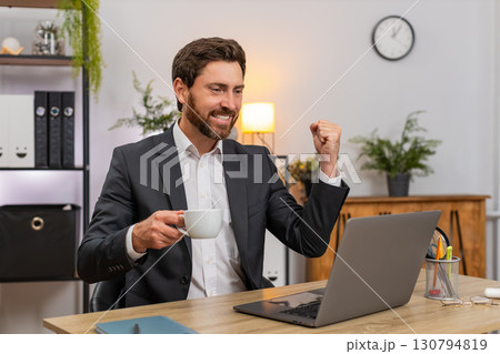 Middle-aged businessman closes laptop raises espresso shot toasts success leans back chair relaxed Middle-aged businessman closes laptop raises espresso shot toasts success leans back chair relaxed 130794819