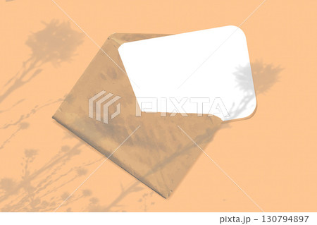 Natural light casts shadows from wildflowers on an envelope with a sheet of white paper lying on an orange background. Mockup 130794897