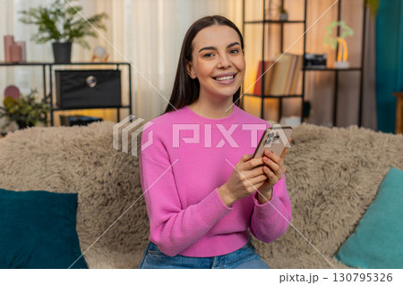 Young woman smiling while browsing social media feed on mobile phone relaxing at cozy home sofa 130795326