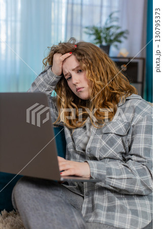 Young woman sits on sofa with laptop upset holding hand on forehead feeling failure and frustration 130795373