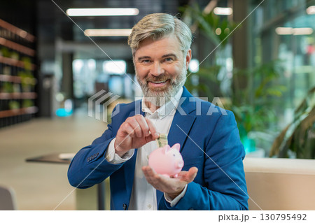 Happy mature businessman save money using pink piggybank putting dollar cast inside in office lobby Happy mature businessman save money using pink piggybank putting dollar cast inside in office lobby 130795492