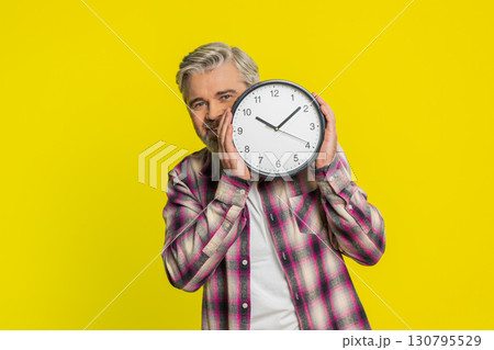 Mature man hiding behind large office clock, playful peek, cautious glance, time awareness humor 130795529