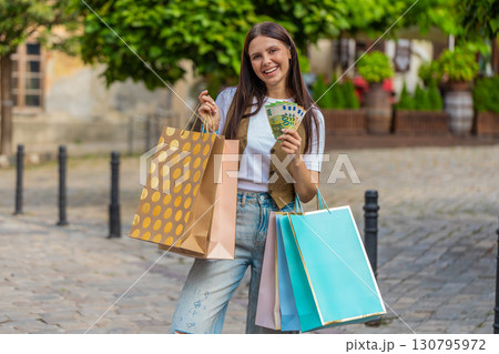 Happy young shopaholic Caucasian woman holding euro cash while carrying shopping bags on city street Happy young shopaholic Caucasian woman holding euro cash while carrying shopping bags on city street 130795972