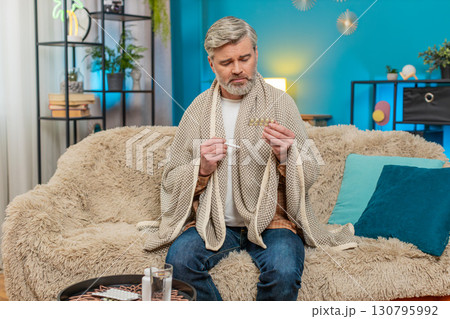 Mature man covered with blanket sitting on sofa, holding thermometer and choosing medicine at home 130795992