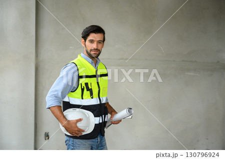 Portrait of young attractive construction man smiling bearded in vest and holding white helmet and blueprint while standing on construction site. Home building concept. 130796924