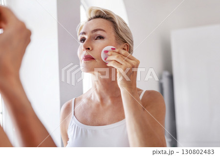 Mature woman 50 years applying skincare with cotton pad, standing by mirror at home. Elegant senior female doing daily beauty routine, natural aging and healthy lifestyle 130802483