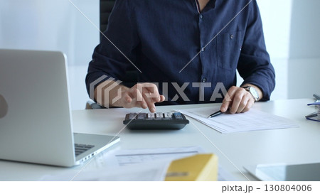 Businessman using a calculator and analyzing financial documents while preparing paperwork at an office desk. Audit and taxes, business concept Businessman using a calculator and analyzing financial documents while preparing paperwork at an office desk. Audit and taxes, business concept 130804006