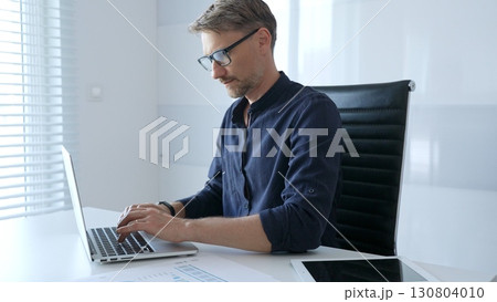 Professional businessman wearing glasses, intensely working on laptop, typing amid modern office environment with tablet and documents surrounding workspace. Business people concept Professional businessman wearing glasses, intensely working on laptop, typing amid modern office environment with tablet and documents surrounding workspace. Business people concept 130804010
