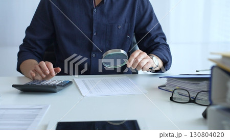 Financial analyst reviewing paperwork, using calculator and magnifying glass while examining corporate budget reports in modern workspace. Audit and taxes, business concept Financial analyst reviewing paperwork, using calculator and magnifying glass while examining corporate budget reports in modern workspace. Audit and taxes, business concept 130804020