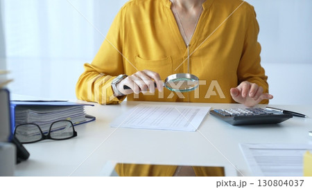 Female accountant wearing yellow shirt is examining a financial report with a magnifying glass while using a calculator and laptop in an office setting, close up. Audit and taxes in business Female accountant wearing yellow shirt is examining a financial report with a magnifying glass while using a calculator and laptop in an office setting, close up. Audit and taxes in business 130804037