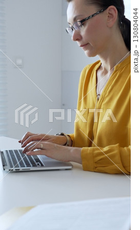 Businesswoman wearing yellow shirt sitting at white desk typing on laptop in bright modern office, working productively on computer. Business people concept 130804044
