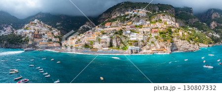 The coastal town of Positano, Italy, features pastel colored buildings on cliffs, a beach with umbrellas, turquoise waters, boats, and lush greenery. 130807332