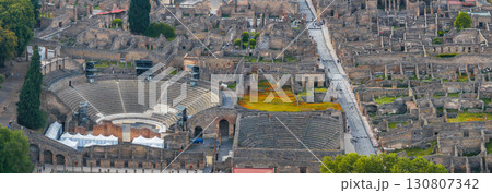 Aerial view of Pompeii ruins in Italy, featuring an amphitheater with semi circular seating, stone buildings, streets, and patches of greenery. 130807342