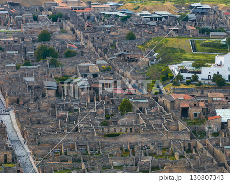 Aerial view of Pompeii's ancient ruins in Italy, showing stone walls, streets, courtyards, and greenery, with modern buildings in the background. 130807343