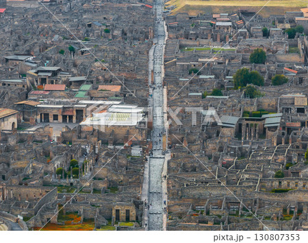Aerial view of Pompeii ruins in Italy, featuring a central stone paved street, grid like city layout, preserved structures, and surrounding greenery. 130807353