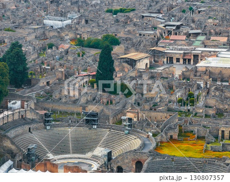 Aerial view of Pompeii's ancient ruins featuring a semi circular stone amphitheater, crumbled buildings, pathways, and greenery with cypress trees. Aerial view of Pompeii's ancient ruins featuring a semi circular stone amphitheater, crumbled buildings, pathways, and greenery with cypress trees. 130807357