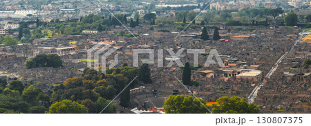 Aerial view of Pompeii's ancient ruins, featuring the amphitheater, stone streets, and buildings, with modern urban areas and greenery in the background. 130807375