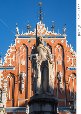A knight statue with a shield stands before The House of the Blackheads in Riga, Latvia, with a blue sky, clouds, and yellow flowers in the foreground. A knight statue with a shield stands before The House of the Blackheads in Riga, Latvia, with a blue sky, clouds, and yellow flowers in the foreground. 130807377