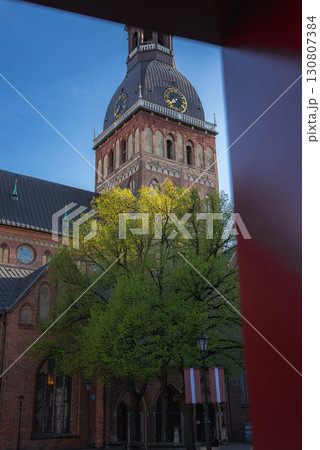 The Riga Cathedral stands with its Gothic architecture and clock tower, framed by a green tree, Latvian flags, a red element, and a bright blue sky. 130807384