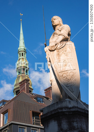 A statue of a historical figure with a sword and shield stands before the spire of St. Peter's Church in Riga, Latvia, under a clear blue sky. 130807400