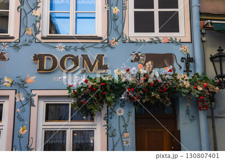 A soft blue building facade in Riga's old town features floral patterns, a whimsical cow illustration, gold DOM sign, and vibrant flower arrangements. 130807401