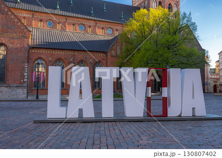 A large LATVIJA installation with a flag inspired J stands in a square near Riga Cathedral. A tree, cobblestone pavement, and blue sky complete the scene. A large LATVIJA installation with a flag inspired J stands in a square near Riga Cathedral. A tree, cobblestone pavement, and blue sky complete the scene. 130807402