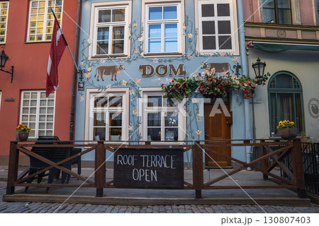 A cafe in Riga's old town features a blue facade with floral and animal motifs, a Latvian flag, outdoor seating, cobblestone street, and nearby buildings. 130807403