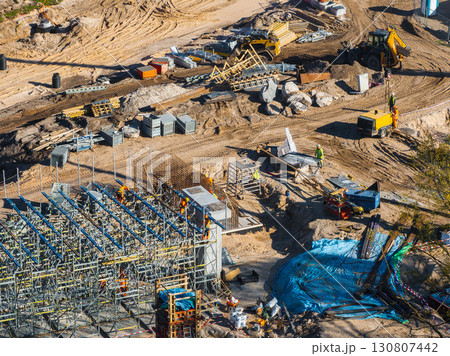 Active construction site with workers, scaffolding, heavy machinery, steel beams, concrete blocks, and a yellow truck in Riga, Latvia. Active construction site with workers, scaffolding, heavy machinery, steel beams, concrete blocks, and a yellow truck in Riga, Latvia. 130807442