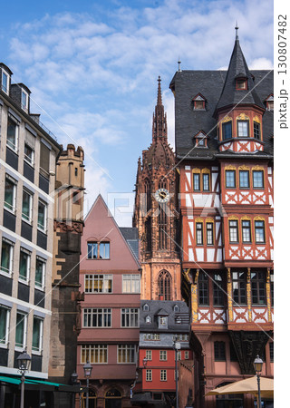 A street in Frankfurt, Germany, featuring the Gothic style Frankfurt Cathedral, a red and white half timbered building, and modern structures under a clear sky. 130807482