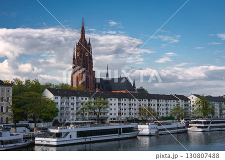 The Frankfurt Cathedral's Gothic spire rises against a partly cloudy sky, with the Main River, riverboats, residential buildings, and greenery in view. The Frankfurt Cathedral's Gothic spire rises against a partly cloudy sky, with the Main River, riverboats, residential buildings, and greenery in view. 130807488