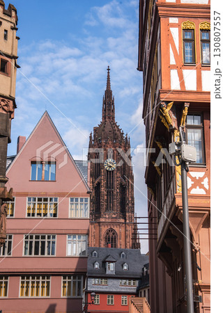 The Gothic style Frankfurt Cathedral stands tall with its intricate spire, surrounded by half timbered and modern buildings under a bright blue sky. 130807557