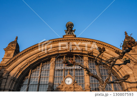 The facade of Frankfurt Hauptbahnhof in Germany features a grand arched window, a central clock, Atlas statue, smaller sculptures, and a clear blue sky. 130807593