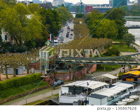 Frankfurt's Main River with a tree lined promenade, docked boats, a pedestrian bridge, urban buildings, and greenery in a vibrant riverside scene. 130807628