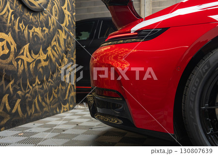 Rear view of a red 2025 Porsche 911 GT3 922 with sleek curves and taillight, parked indoors on a checkered floor with graffiti style wall behind. 130807639
