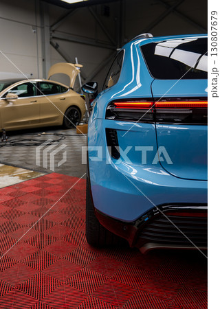 Rear view of a blue Porsche Macan on a red floor mat in a garage. A beige car with its hood open and tools are visible in the industrial space. Rear view of a blue Porsche Macan on a red floor mat in a garage. A beige car with its hood open and tools are visible in the industrial space. 130807679