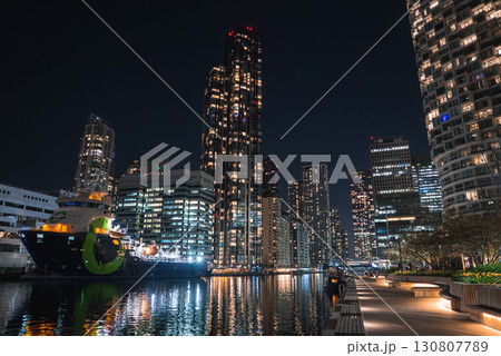 Canary Wharf's illuminated skyscrapers at night, a green hulled docked ship, calm canal waters, a lit boardwalk, benches, and landscaped greenery. 130807789