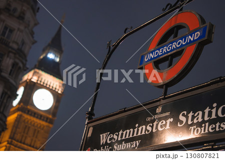 The Westminster Station sign with the red and blue roundel logo stands in the foreground, while Big Ben's illuminated clock face glows in the night sky. The Westminster Station sign with the red and blue roundel logo stands in the foreground, while Big Ben's illuminated clock face glows in the night sky. 130807821