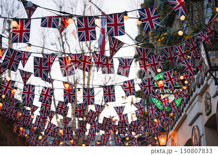 A London street decorated with Union Jack flags and warm string lights. Traditional architecture, bare trees, and greenery enhance the urban scene. 130807833