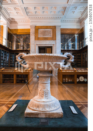 A white marble urn on a green pedestal in a library with wooden display cases, glass enclosed bookshelves, and a coffered ceiling in London. A white marble urn on a green pedestal in a library with wooden display cases, glass enclosed bookshelves, and a coffered ceiling in London. 130807838