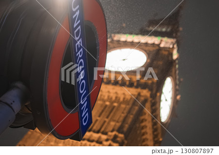 The iconic London Underground sign in the foreground with Big Ben's illuminated clock face in the background, captured at night with a soft glow and mist. 130807897