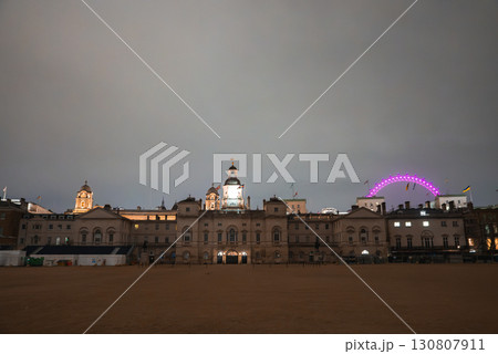 Horse Guards Parade with its clock tower lit against the evening sky, the London Eye glowing pink to the right, and an empty sandy parade ground. Horse Guards Parade with its clock tower lit against the evening sky, the London Eye glowing pink to the right, and an empty sandy parade ground. 130807911